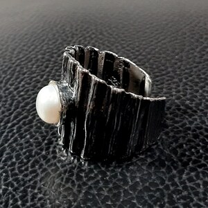May include: A silver ring with a textured, wood-like band and a white pearl set in the front.