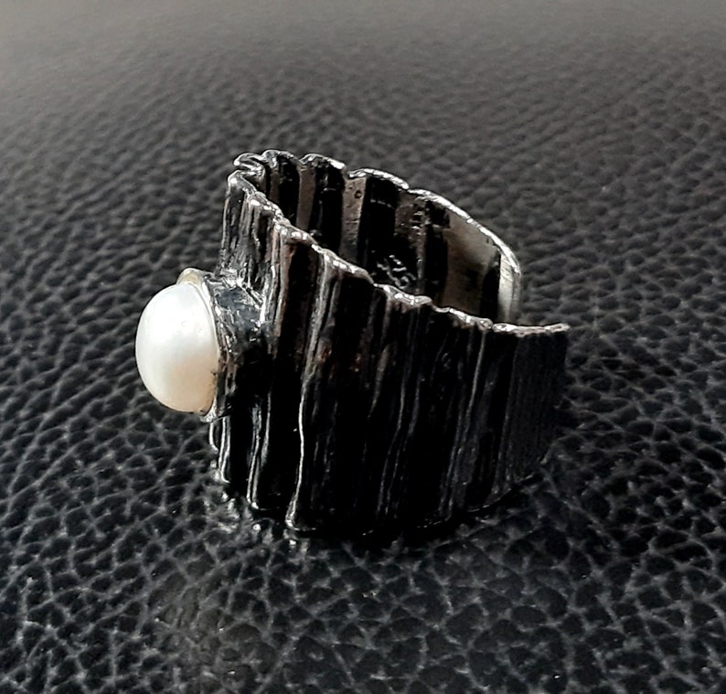 May include: A silver ring with a textured, wood-like band and a white pearl set in the front.