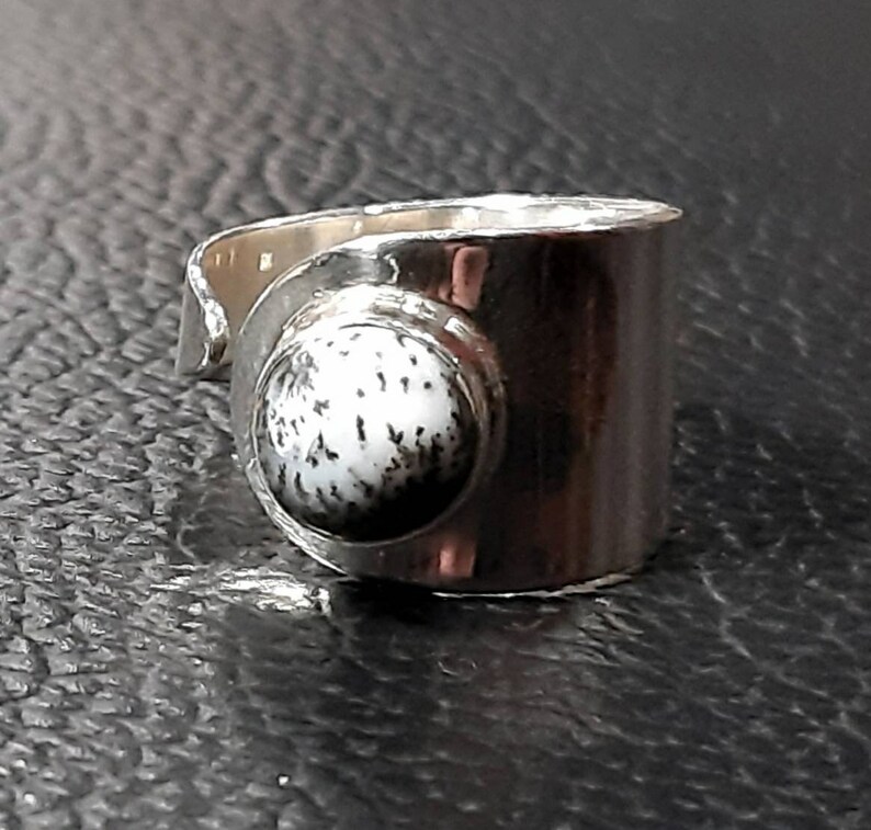 May include: A silver ring with a white and black stone set in the center. The ring is wide and has a simple design.