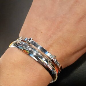 May include: Two silver bangle bracelets on a person's wrist. The bracelets are both smooth and shiny, and one has a small diamond-like accent.