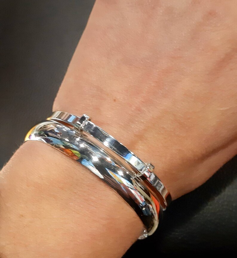 May include: Two silver bangle bracelets on a person's wrist. The bracelets are both smooth and shiny, and one has a small diamond-like accent.