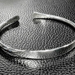 May include: A silver bangle bracelet with a smooth, rounded surface. The bracelet is open and has a simple, minimalist design.