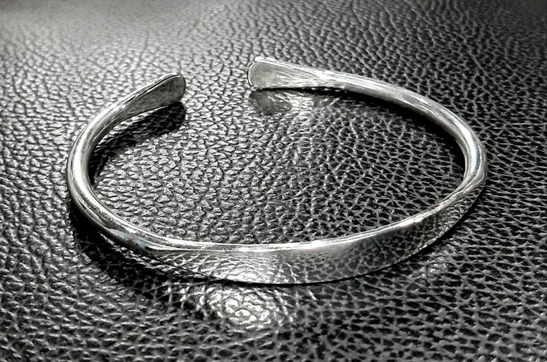 May include: A silver bangle bracelet with a smooth, rounded surface. The bracelet is open and has a simple, minimalist design.