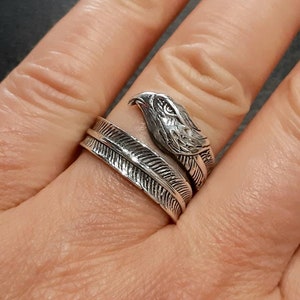 May include: A silver-colored ring featuring an eagle head design. The ring wraps around the finger with a feather motif. The eagle's head is detailed with textured feathers and a sharp beak. The ring is worn on a finger.