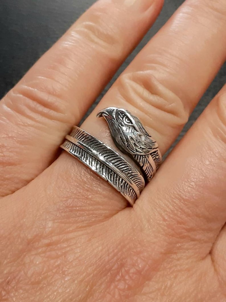 May include: A silver-colored ring featuring an eagle head design. The ring wraps around the finger with a feather motif. The eagle's head is detailed with textured feathers and a sharp beak. The ring is worn on a finger.