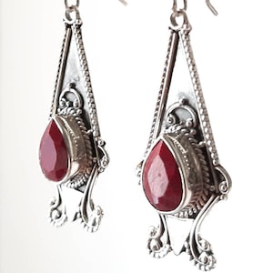May include: A pair of silver dangle earrings with red gemstone drops. The earrings feature intricate silver designs and a teardrop-shaped red gemstone.