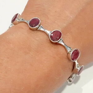 May include: A silver bracelet with a series of oval-shaped red gemstones. The bracelet is open and has a hinged clasp.