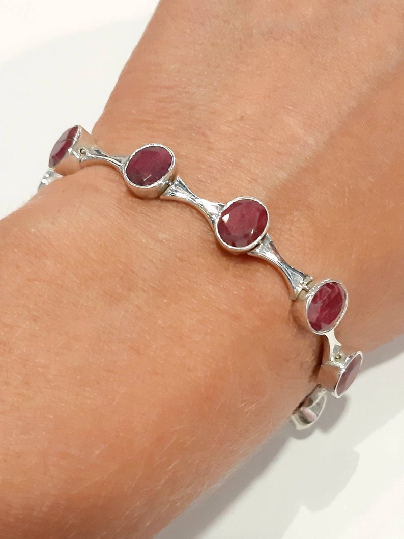 May include: A silver bracelet with a series of oval-shaped red gemstones. The bracelet is open and has a hinged clasp.
