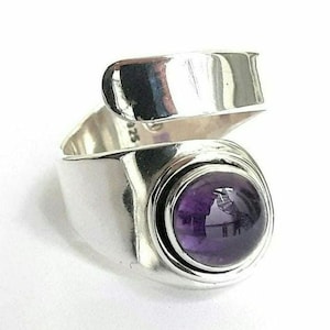 Pode incluir: Um anel de estilo bypass feito de prata esterlina polida, com uma banda larga e lisa que se sobrepõe no topo. Uma única pedra preciosa de ametista roxa, redonda e lapidada em cabochão, está engastada na frente, com a marca 925 visível no interior.