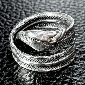 May include: A silver-colored ring featuring an eagle head design. The ring wraps around the finger with a feather-like pattern. The eagle's head is detailed with a sharp beak and focused gaze. The ring is made of a shiny metal.