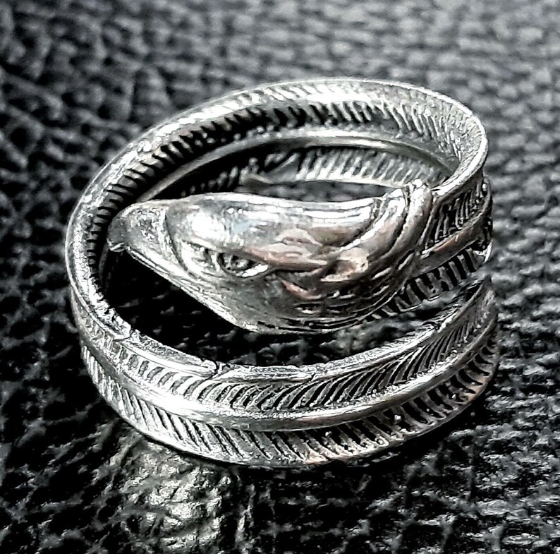 May include: A silver-colored ring featuring an eagle head design. The ring wraps around the finger with a feather-like pattern. The eagle's head is detailed with a sharp beak and focused gaze. The ring is made of a shiny metal.