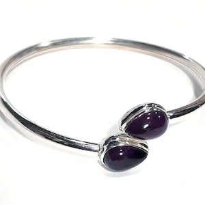 Peut inclure: Un bracelet jonc en argent avec deux accents de pierres précieuses violettes. Le bracelet est ouvert et a un design simple.