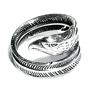 May include: A silver-colored ring featuring an eagle head design. The ring wraps around the finger with a feather-like pattern on the band. The eagle's head is detailed with a sharp beak and eye.