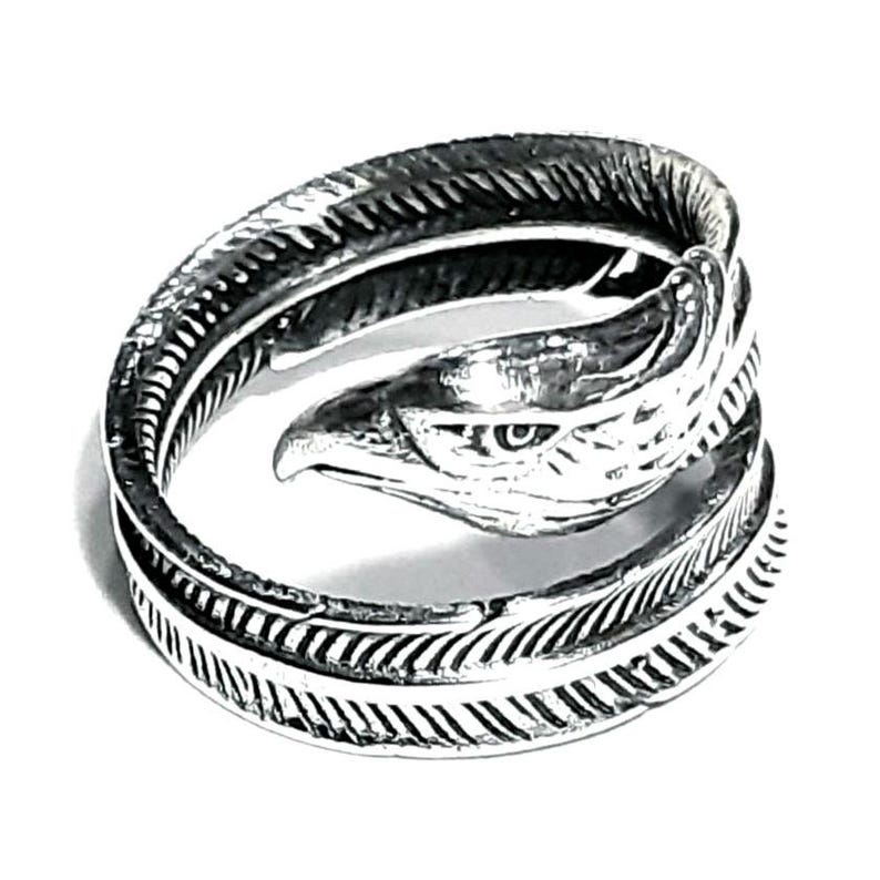 May include: A silver-colored ring featuring an eagle head design. The ring wraps around the finger with a feather-like pattern on the band. The eagle's head is detailed with a sharp beak and eye.