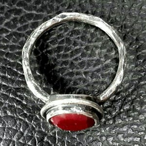 Pu&ograve; includere: Un anello d'argento con una pietra preziosa rossa incastonata in una montatura a castone. La fascia dell'anello &egrave; testurizzata e ha una finitura martellata.