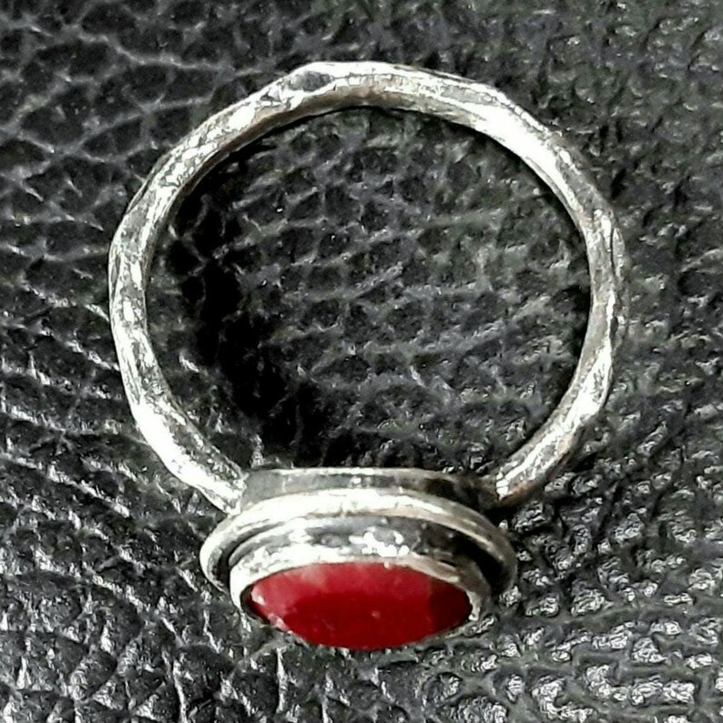 Pu&ograve; includere: Un anello d'argento con una pietra preziosa rossa incastonata in una montatura a castone. La fascia dell'anello &egrave; testurizzata e ha una finitura martellata.