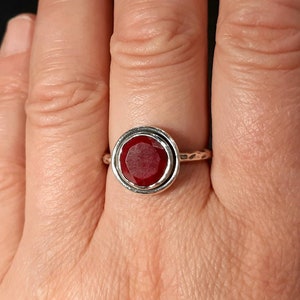 Pu&ograve; includere: Un anello d'argento con una pietra preziosa rossa incastonata in una montatura a bezel. L'anello ha una fascia testurizzata.
