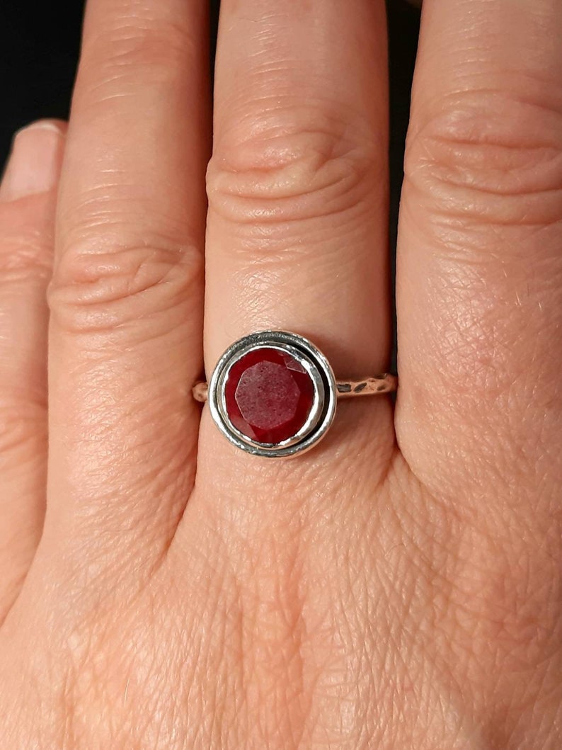 Pu&ograve; includere: Un anello d'argento con una pietra preziosa rossa incastonata in una montatura a bezel. L'anello ha una fascia testurizzata.