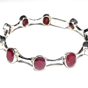 May include: A silver bangle bracelet with red gemstones. The bracelet is made of a series of connected silver links, each with a red gemstone set in the center.