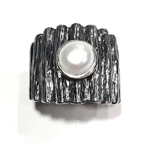 May include: A silver ring with a textured, wood-like band and a white pearl set in the center.