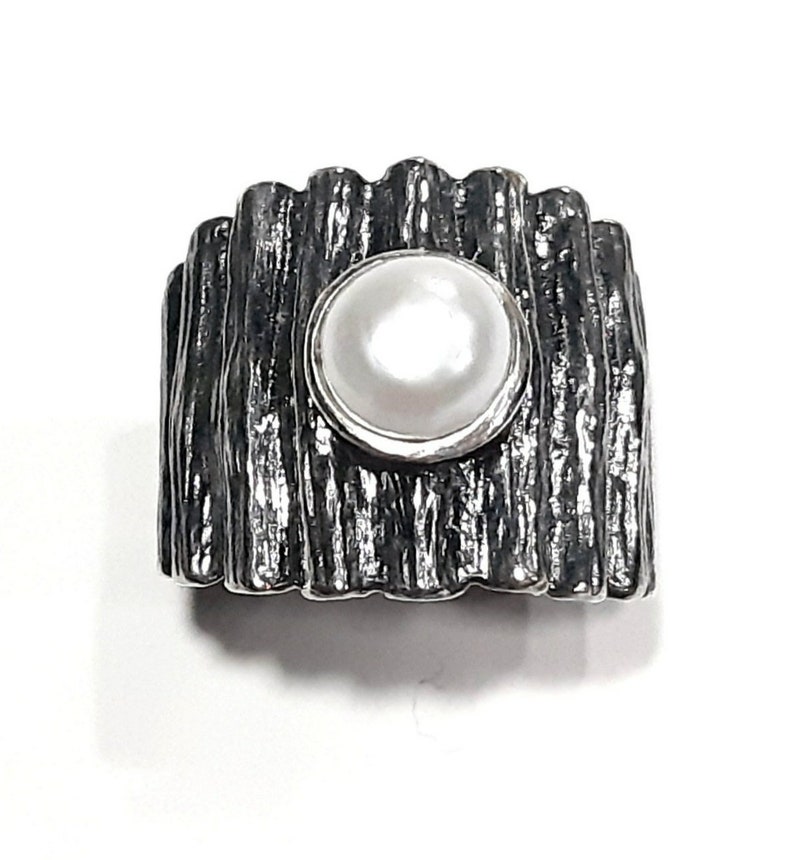 May include: A silver ring with a textured, wood-like band and a white pearl set in the center.