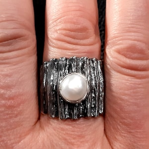 May include: A silver ring with a textured band and a white pearl in the center.