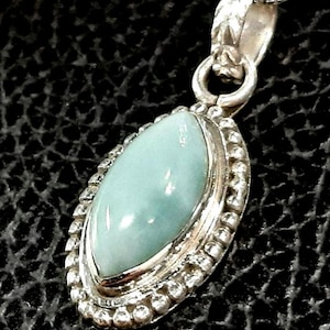 May include: A silver pendant necklace with a light blue gemstone set in a silver bezel. The pendant is surrounded by a beaded border.