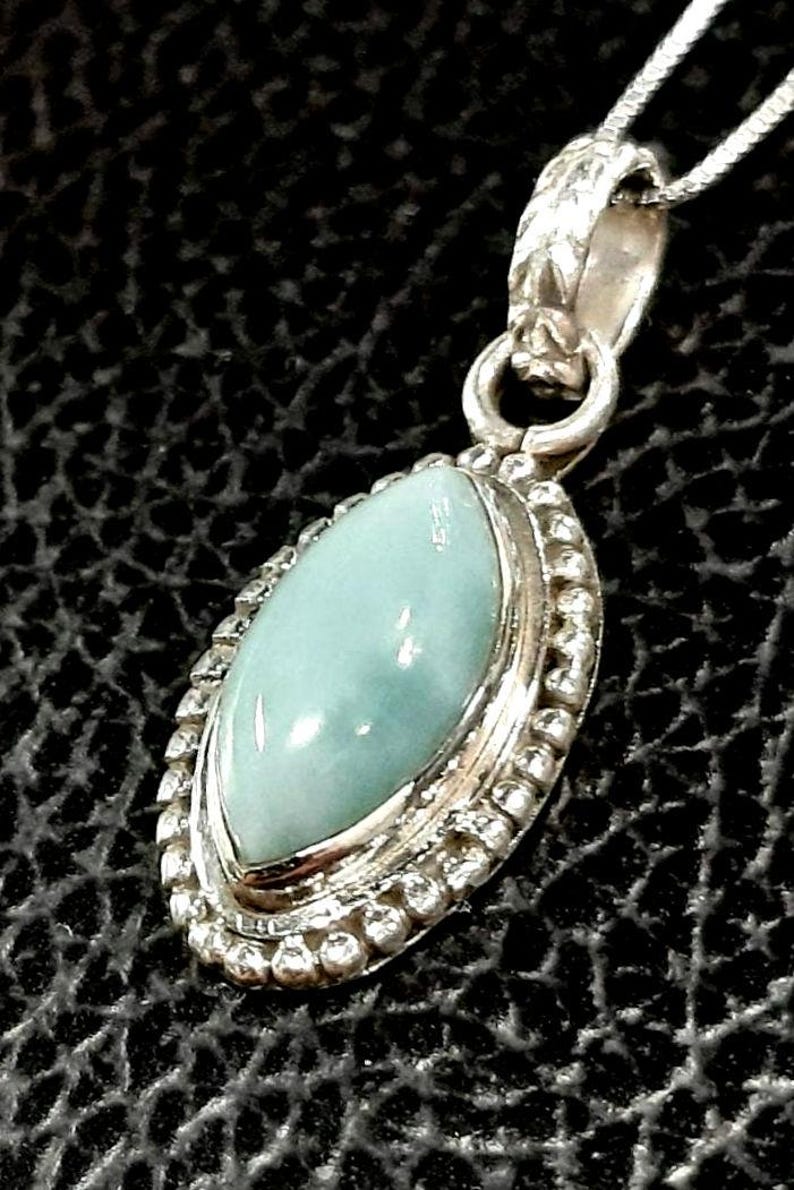 May include: A silver pendant necklace with a light blue gemstone set in a silver bezel. The pendant is surrounded by a beaded border.
