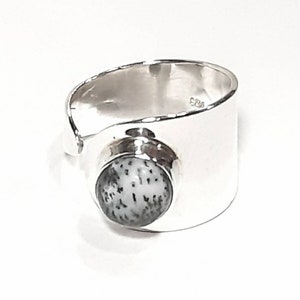 May include: A silver ring with a white and gray stone set in the center. The ring is wide and has a smooth, polished finish.