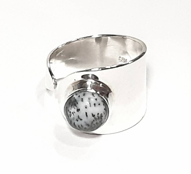 May include: A silver ring with a white and gray stone set in the center. The ring is wide and has a smooth, polished finish.