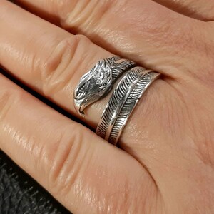 May include: A silver-colored ring featuring an eagle head design. The ring wraps around the finger with a feather-like band. The eagle head has detailed features, and the ring is worn on a hand.