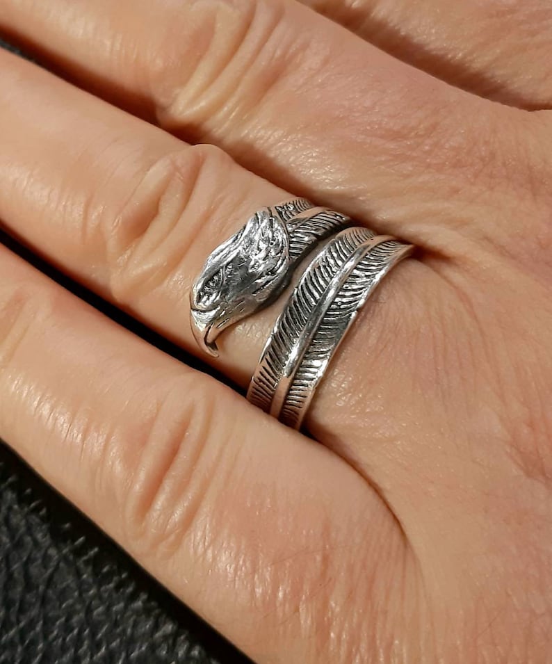 May include: A silver-colored ring featuring an eagle head design. The ring wraps around the finger with a feather-like band. The eagle head has detailed features, and the ring is worn on a hand.
