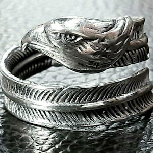 May include: A silver-toned ring featuring an eagle head design and a feather motif. The eagle's head is detailed with sharp features, and the feather wraps around the finger. The ring is made of a shiny metal.