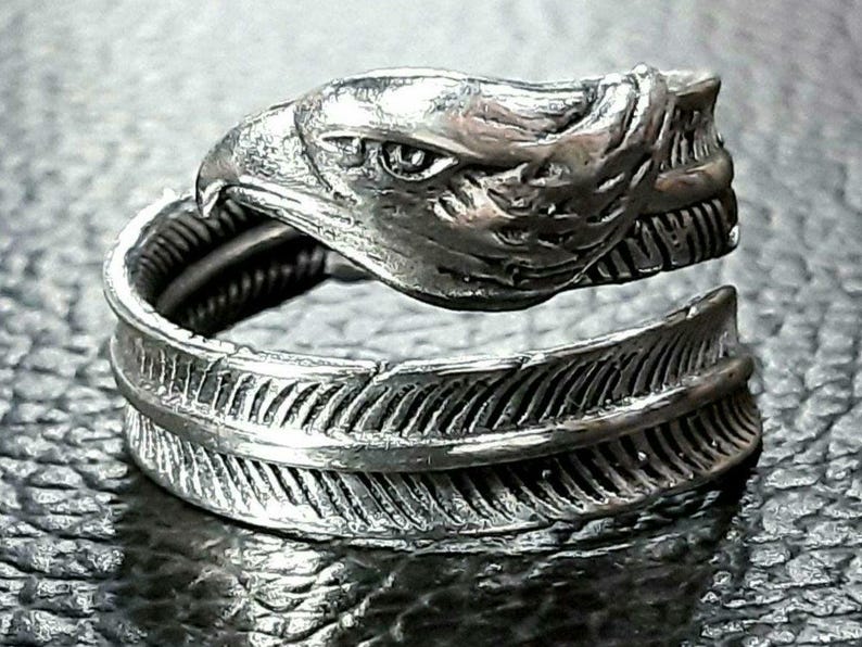 May include: A silver-toned ring featuring an eagle head design and a feather motif. The eagle's head is detailed with sharp features, and the feather wraps around the finger. The ring is made of a shiny metal.