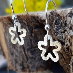 K&ouml;nnte beinhalten: Silberne Blumenohrringe mit mattem Finish. Die Ohrringe haben ein kleines Blumendesign mit einem ausgeschnittenen Mittelteil, das an einem einfachen Haken h&auml;ngt. Der Hintergrund umfasst eine Holzoberfl&auml;che und gelbe Blumen.