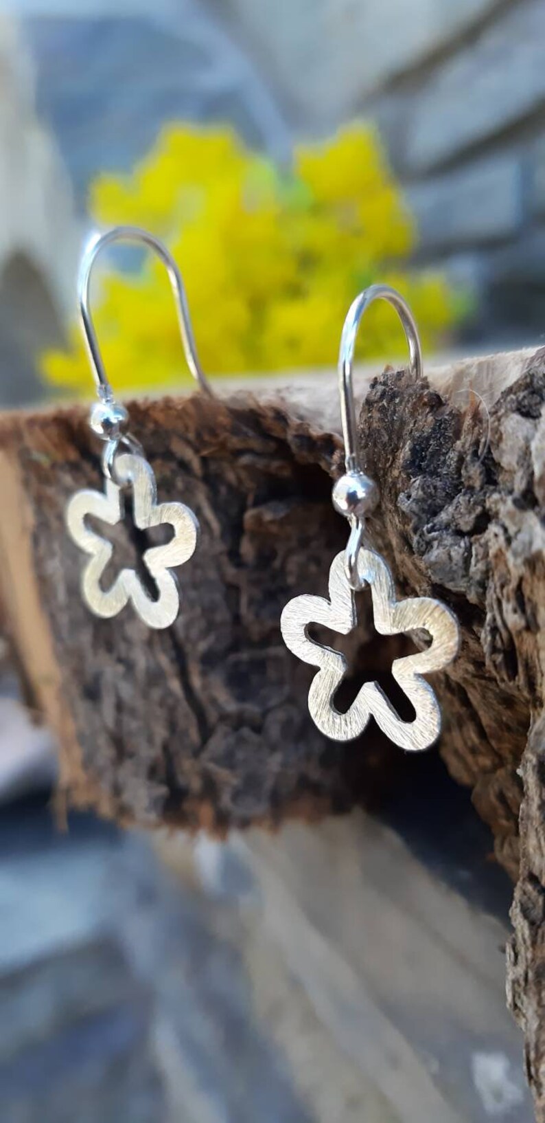 K&ouml;nnte beinhalten: Silberne Blumenohrringe mit mattem Finish. Die Ohrringe haben ein kleines Blumendesign mit einem ausgeschnittenen Mittelteil, das an einem einfachen Haken h&auml;ngt. Der Hintergrund umfasst eine Holzoberfl&auml;che und gelbe Blumen.