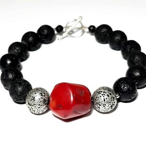 Puede incluir: Un brazalete de cuentas de roca volc&aacute;nica negra con una cuenta de coral rojo y dos cuentas plateadas. El brazalete tiene un cierre de palanca plateado.
