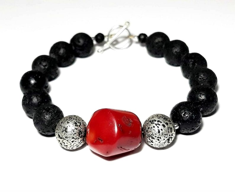 Puede incluir: Un brazalete de cuentas de roca volc&aacute;nica negra con una cuenta de coral rojo y dos cuentas plateadas. El brazalete tiene un cierre de palanca plateado.