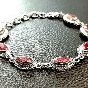 May include: A silver bracelet with red gemstones set in oval-shaped bezels. The bracelet has a delicate chain link design and is adorned with intricate details.