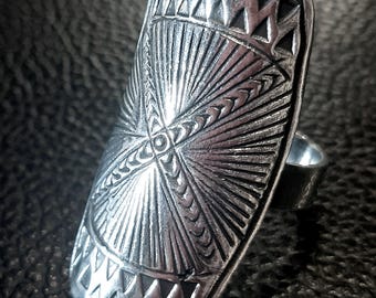May include: A silver-toned ring with an elongated rectangular face. The face is intricately detailed with geometric patterns, including triangles and radiating lines. The ring has a polished finish and a simple band.