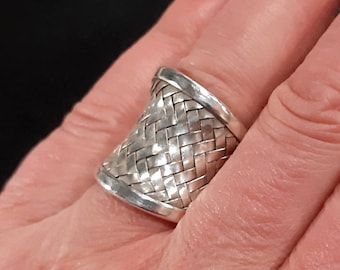 May include: A silver ring with a woven basket design. The ring has a wide band with a textured pattern, and a smooth, polished finish. The ring is a statement piece, suitable for everyday wear or special occasions.