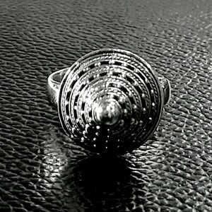 May include: A silver ring with a circular design featuring concentric circles and a raised center. The ring is made of a textured metal and has a geometric pattern.