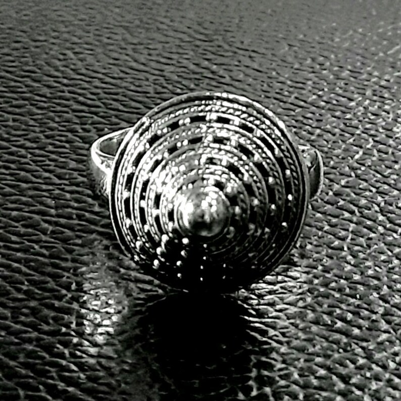 May include: A silver ring with a circular design featuring concentric circles and a raised center. The ring is made of a textured metal and has a geometric pattern.