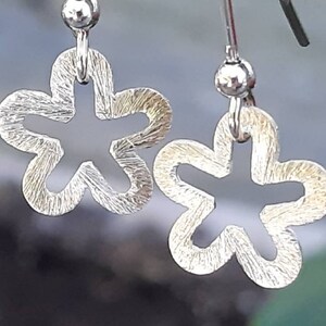 K&ouml;nnte beinhalten: Ein Paar silberfarbene Ohrringe in Blumenform. Jeder Ohrring hat eine strukturierte Oberfl&auml;che und einen h&auml;ngenden Haken. Die Ohrringe sind mit einer f&uuml;nfbl&auml;ttrigen Bl&uuml;tenform mit einem ausgeschnittenen Mittelteil gestaltet.