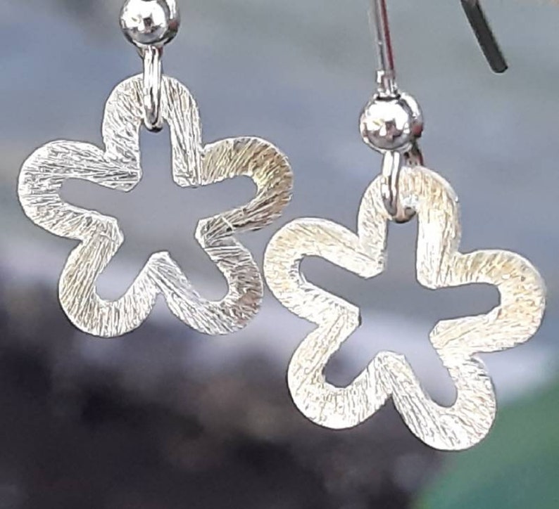 K&ouml;nnte beinhalten: Ein Paar silberfarbene Ohrringe in Blumenform. Jeder Ohrring hat eine strukturierte Oberfl&auml;che und einen h&auml;ngenden Haken. Die Ohrringe sind mit einer f&uuml;nfbl&auml;ttrigen Bl&uuml;tenform mit einem ausgeschnittenen Mittelteil gestaltet.