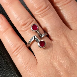 Op de afbeelding: Een zilveren ring met twee rode edelstenen. De ring heeft een geometrisch ontwerp en is getextureerd.