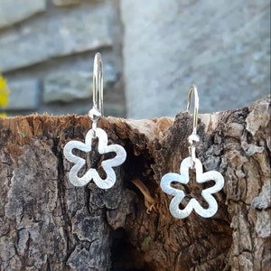 K&ouml;nnte beinhalten: Ein Paar silberne, bl&uuml;tenf&ouml;rmige Ohrh&auml;nger. Jeder Ohrring hat ein f&uuml;nfbl&auml;ttriges Blumendesign mit strukturierter Oberfl&auml;che, das an einem einfachen Haken h&auml;ngt. Die Ohrringe werden auf einem St&uuml;ck strukturiertem Holz pr&auml;sentiert.