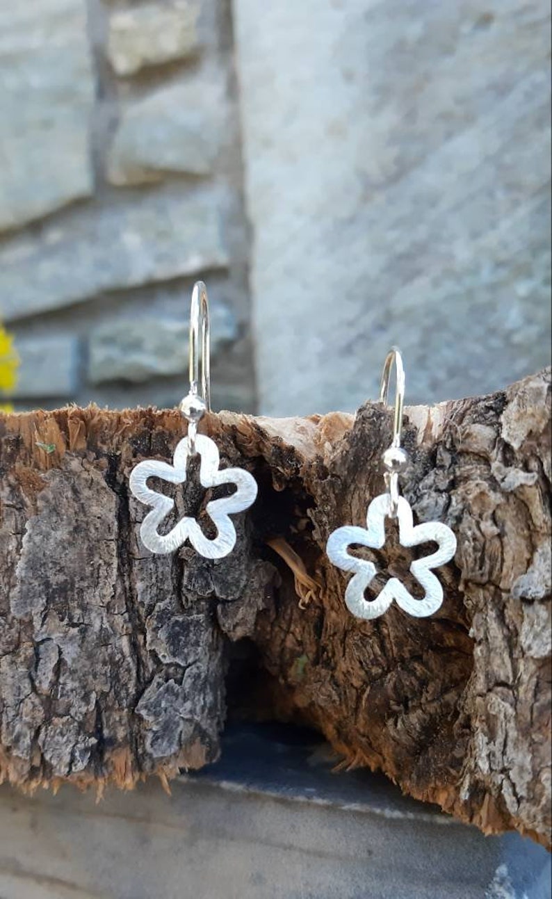 K&ouml;nnte beinhalten: Ein Paar silberne, bl&uuml;tenf&ouml;rmige Ohrh&auml;nger. Jeder Ohrring hat ein f&uuml;nfbl&auml;ttriges Blumendesign mit strukturierter Oberfl&auml;che, das an einem einfachen Haken h&auml;ngt. Die Ohrringe werden auf einem St&uuml;ck strukturiertem Holz pr&auml;sentiert.