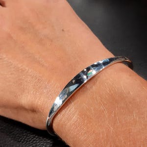 May include: A silver bangle bracelet with a smooth, rounded surface. The bracelet is open and has a slight curve to it.