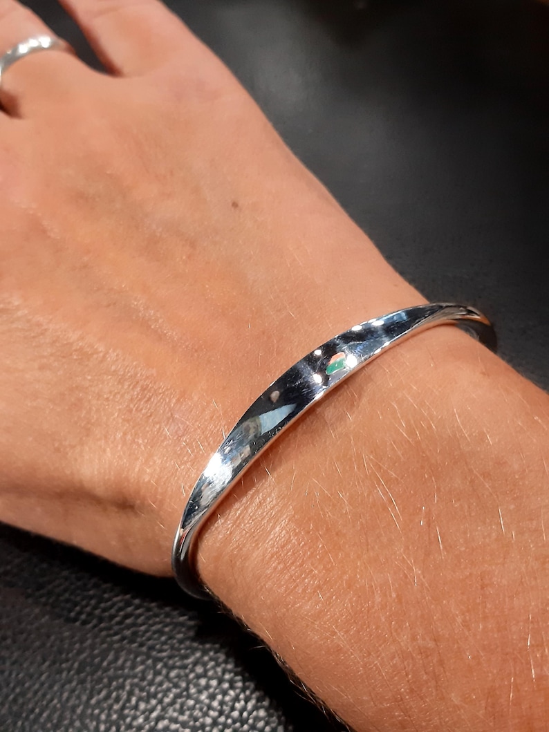 May include: A silver bangle bracelet with a smooth, rounded surface. The bracelet is open and has a slight curve to it.